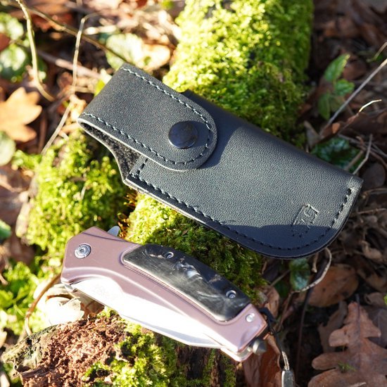 Zwart leren hoes met drukknop en inklapbaar mes met houten handvat op mosrijke omgeving