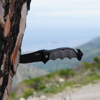 Zwart inklapbaar mes met metalen handvat gepositioneerd in een boomstam in natuurgebied.
