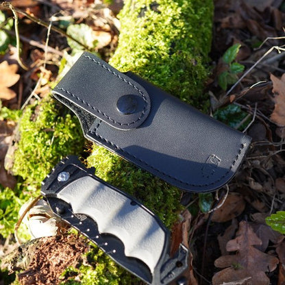 Zwart leren hoesje met drukknop naast zwart-witte vouwmes op mosrijke bosgrond