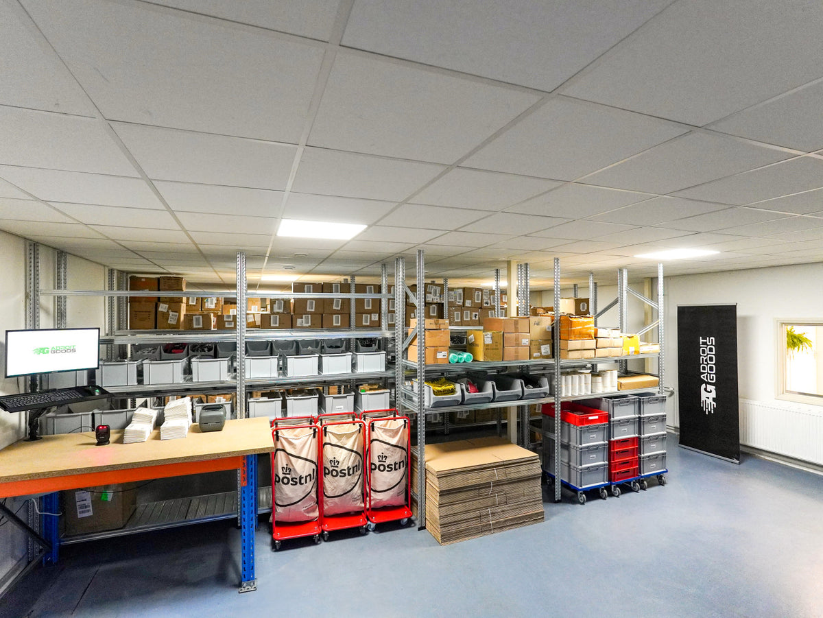 Overzicht van een magazijn met stellingen vol dozen en posttassen bij werktafels in een nette opslagruimte.