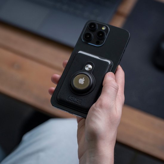 Hand houdt zwarte smartphone met leren portemonnee en AirTaghouder aan achterkant vast