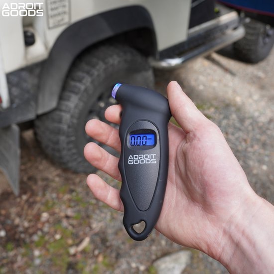 Hand houdt een zwart digitaal apparaat met blauwe display voor het meten van bandenspanning naast een terreinwagenband.