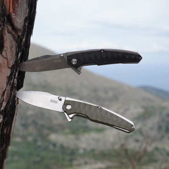 Twee inklapbare messen, één zwart en één zilver, geplaatst in een boomstam met een berglandschap op de achtergrond