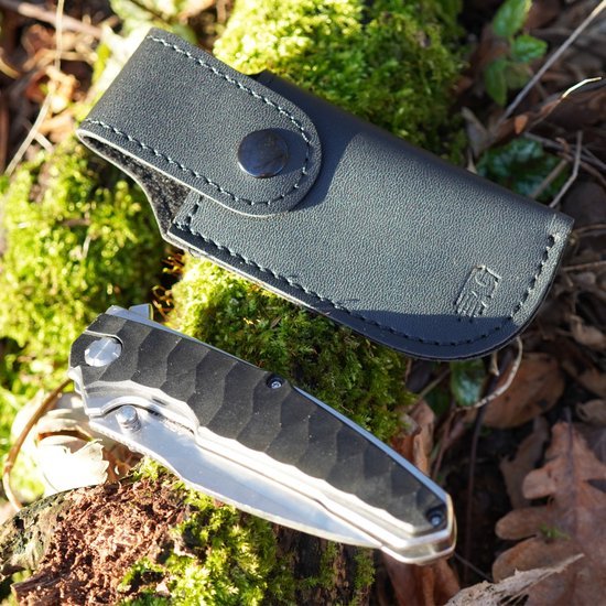 Vouwmes met metalen handgreep en zwart leren hoesje op mos en bladeren in buitenomgeving