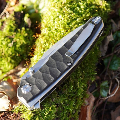 Gesloten zakmes met zwarte, getextureerde handgreep liggend op groene mosbodem buiten.