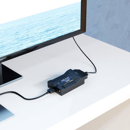 Zwart elektronisch apparaat verbonden met een televisie op een witte tafel in een lichte kamer.