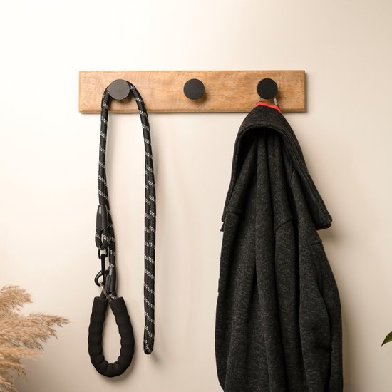 Houten kapstok met zwarte haak, waarop een riem en een donkergrijze jas hangen aan een witte muur.