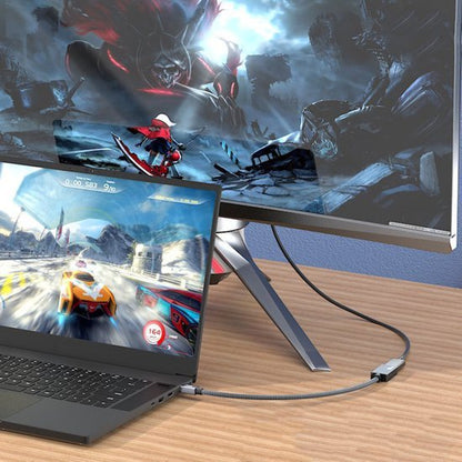 Laptop verbonden met een computerscherm dat actievolle gamebeelden toont op een houten bureau