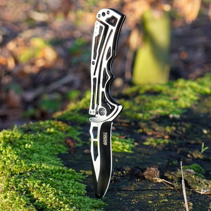 Zilverkleurig inklapbaar mes met zwart lemmet, staand in mosrijke bosgrond.