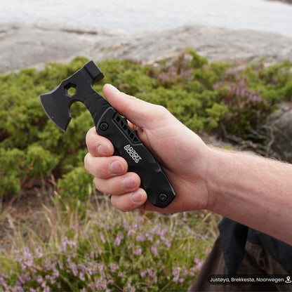 Hand die een compact zwart handbijltje vasthoudt in een groene buitenomgeving