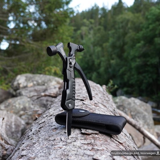 Zwart multifunctioneel multitool met messen en tang op een boomstam in bosrijke omgeving.