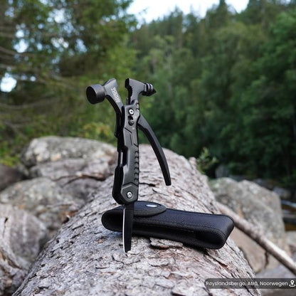 Zwart multifunctioneel multitool met messen en tang op een boomstam in bosrijke omgeving.