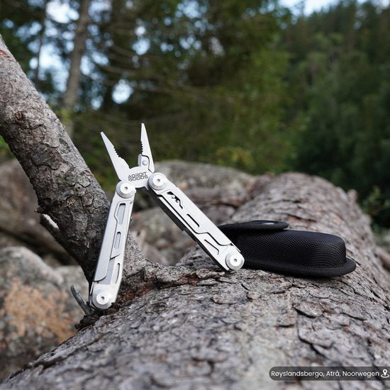 Zilverkleurige multitool geopend op boomstam met zwarte hoes in bosrijke omgeving.