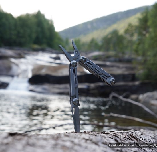 Multitool met tang en mes, geplaatst op een rots bij een stroom in een bosrijke omgeving.