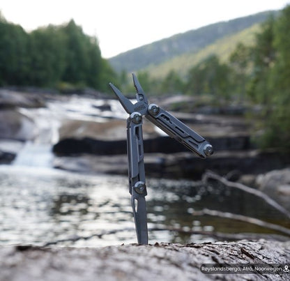 Multitool met tang en mes, geplaatst op een rots bij een stroom in een bosrijke omgeving.