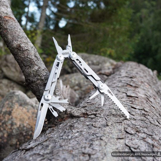 RVS multitool met diverse functies, geplaatst op boomstam in bosrijke omgeving.