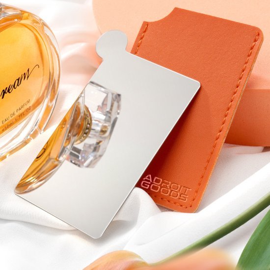 gladde rechthoekige handspiegel met oranje hoes op witte stof naast parfumfles