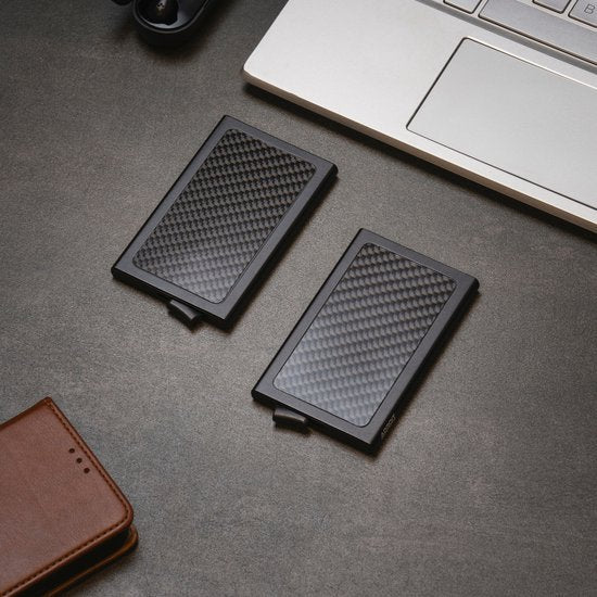 Twee zwarte carbon-look kaartcases liggen naast een laptop en een leren portemonnee op een grijze tafel.