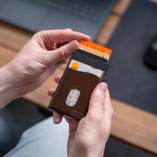 Hand houdt een slanke bruine portemonnee met meerdere kaarten voor een laptop.