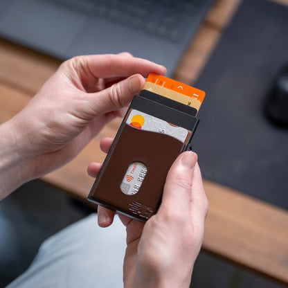 Hand houdt een slanke bruine portemonnee met meerdere kaarten voor een laptop.
