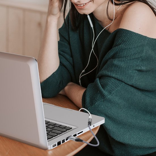 Vrouw met groene trui luistert naar muziek via oordopjes aangesloten op een laptop aan tafel.