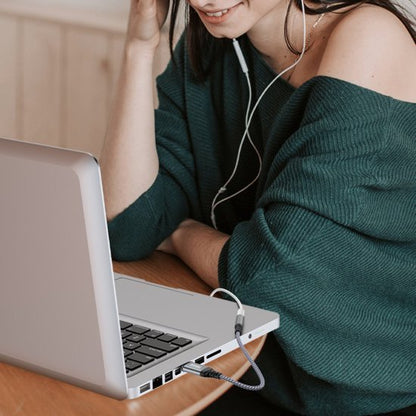 Vrouw met groene trui luistert naar muziek via oordopjes aangesloten op een laptop aan tafel.