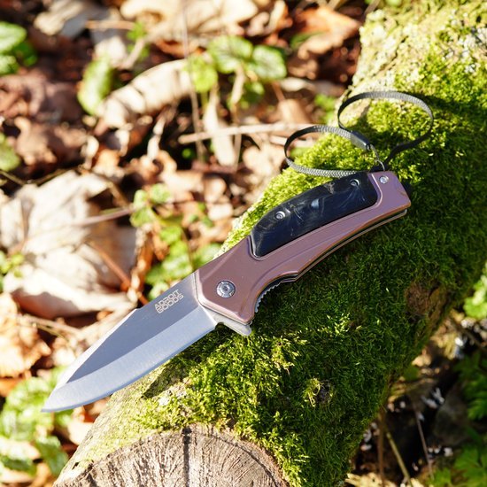 Opvouwbaar mes met zwarte handgreep en messingkleurige details liggend op een mosbedekt hout