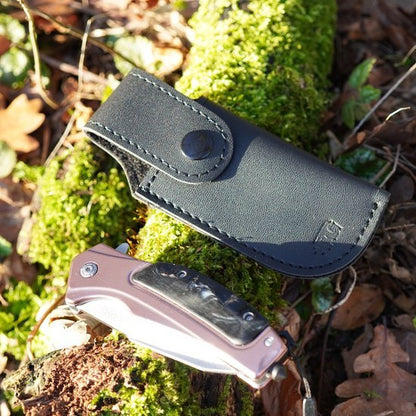 Zwart leren hoes met drukknop en inklapbaar mes met houten handvat op mosrijke omgeving