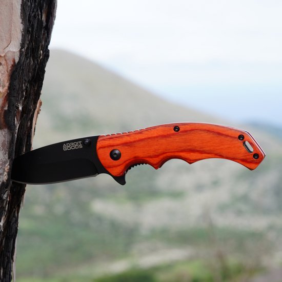 Opvouwbaar mes met zwarte kling en houten handvat bevestigd aan boomstam in natuurlijke omgeving