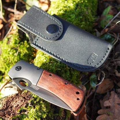 Vouwmes met houten handgreep en leren hoes ligt op mos en bladeren in de natuuromgeving.