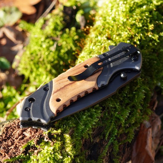 Opvouwbaar mes met houten handvat en zwarte metalen clip liggend op mos en hout