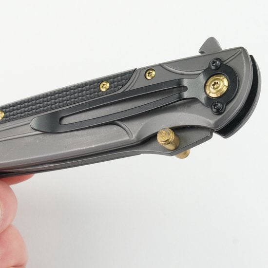 Hand houdt een zwartgrijs inklapbaar mes met gouden schroeven en clip vast tegen een witte achtergrond.