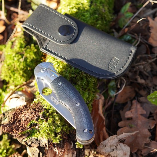 Opgevouwen zakmes met zwarte handgreep naast zwarte leren hoes op mos en herfstbladeren