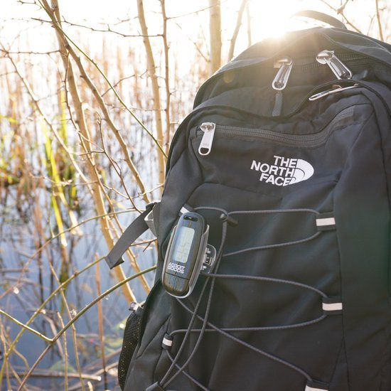 Zwarte rugzak met logo in zonlicht, bevestigd apparaat en elastische koorden buiten in de natuur