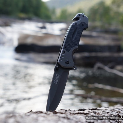 Inklapbaar mes met zwart handvat, in de grond geplaatst bij een rivierlandschap.