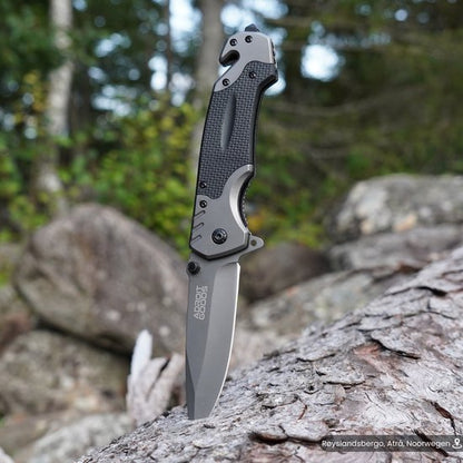 Vouwmes met zwart grip handvat en grijze lemmet, geplaatst op boomstam in bosrijke omgeving