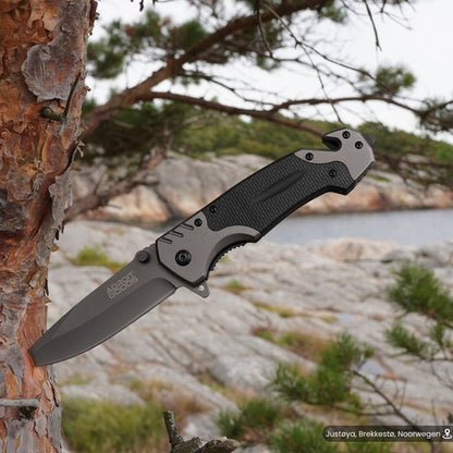 Opgevouwen zakmes met zwart handvat en grijze lemmet in een natuurlandschap met bomen.