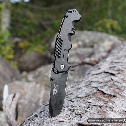 Op tafel geplaatste zwarte vouwmes met robuust handvat en scherp lemmet in bosrijke omgeving