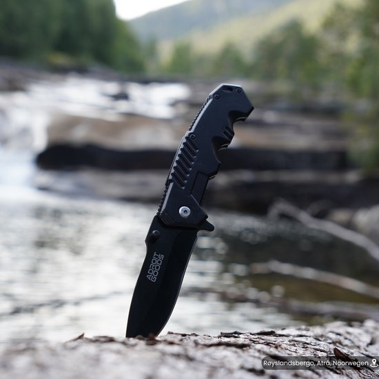 Zwart inklapbaar mes met AdroitGoods logo in natuurlijke omgeving bij stromend water.