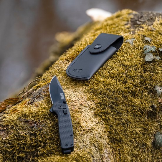 Opgevouwen zakmes met zwarte handgreep en leren hoes op mos bedekt hout in natuurgebied.