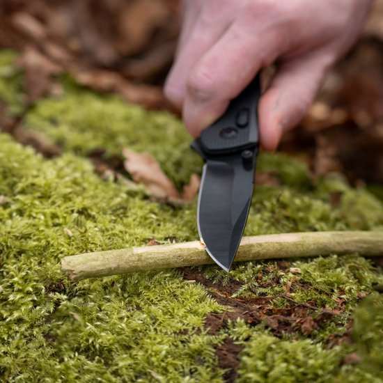 Hand snijdt dun takje met zwart mes op groene mosachtige ondergrond in buitenomgeving