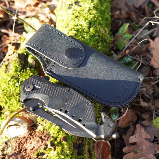 Zwart inklapbaar zakmes met ergonomische grip ligt op mos met leren hoes ernaast geplaatst.