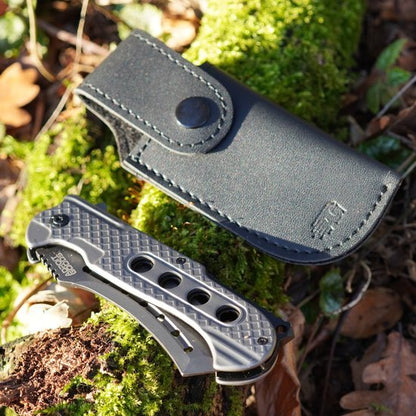 Opvouwbaar metalen zakmes met gegraveerd ontwerp naast een zwarte leren hoes op mosgrond
