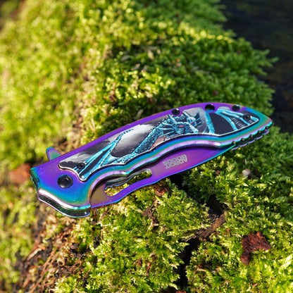 Gesloten kleurig zakmes met drakentekening liggend op mosgroene ondergrond in zonlicht.