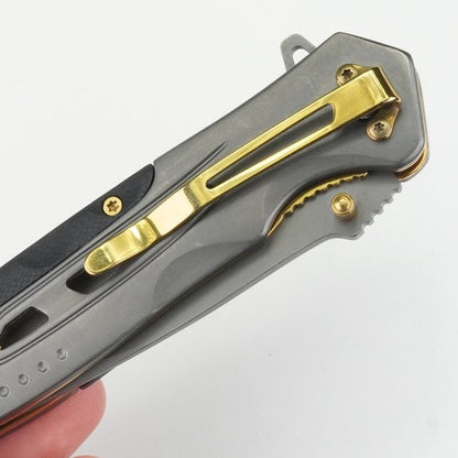 Close-up van een grijs metalen vouwmes met een goudkleurige clip en schroeven in een hand