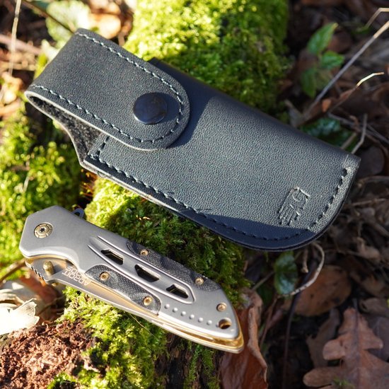 Opgevouwen zilveren multitool met lederen hoes op mosbedekte bosgrond geplaatst