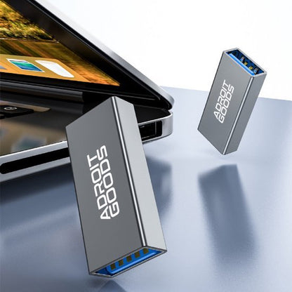 Twee zilveren USB 3.0 adapters met AdroitGoods-logo naast een laptoppoort op een tafelblad.