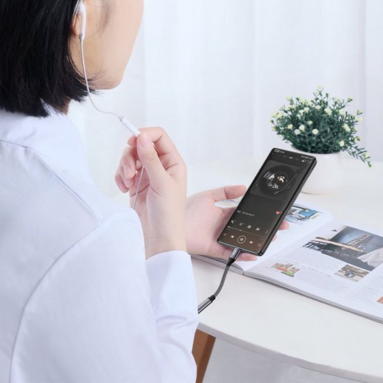 Vrouw met witte oordopjes die naar muziek op een smartphone luistert aan een bureau met plantje en tijdschrift.