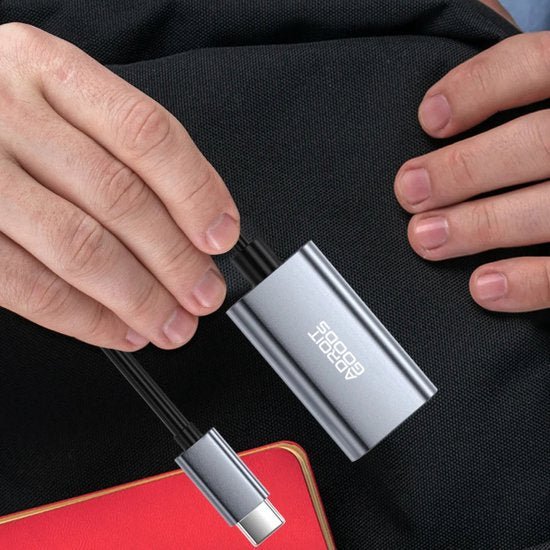 Handen die een compacte grijze adapter met USB-C-kabel op een zwarte ondergrond vasthouden.