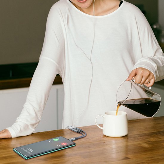 Persoon in witte trui schenkt koffie in een mok naast smartphone met oordopjes op tafel